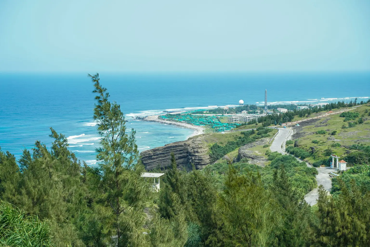 A blue coastline seen from a wooded hillside with a rock cliff, a hillside road and green grass