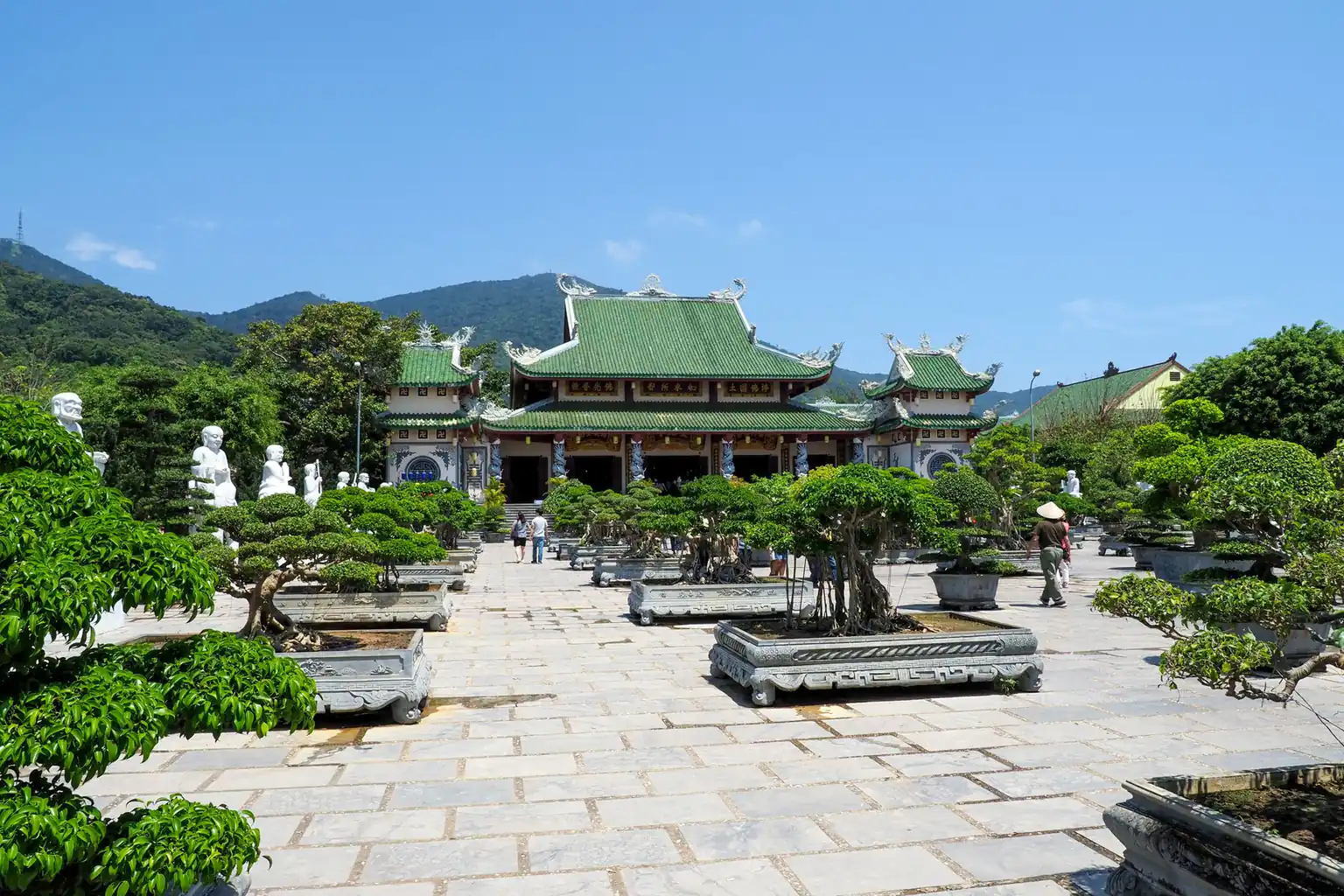 A green-roofed temple hall seen across a stone plaza lined with bonsai trees
