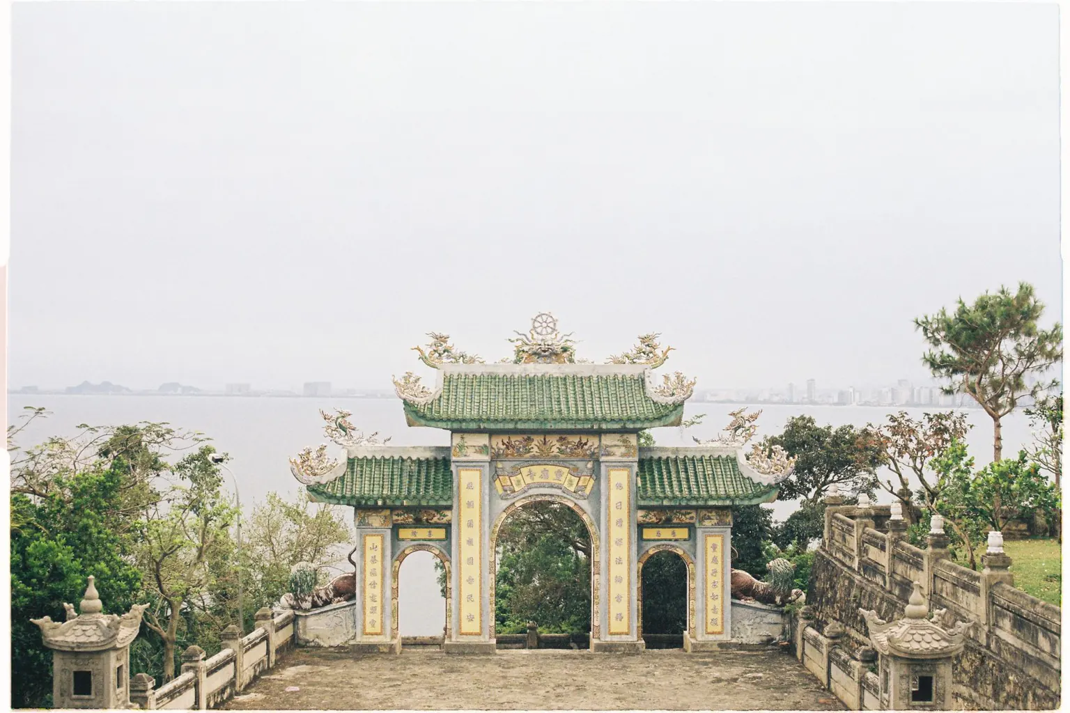 A green-roofed temple gate on a hillside overlooking a hazy sea
