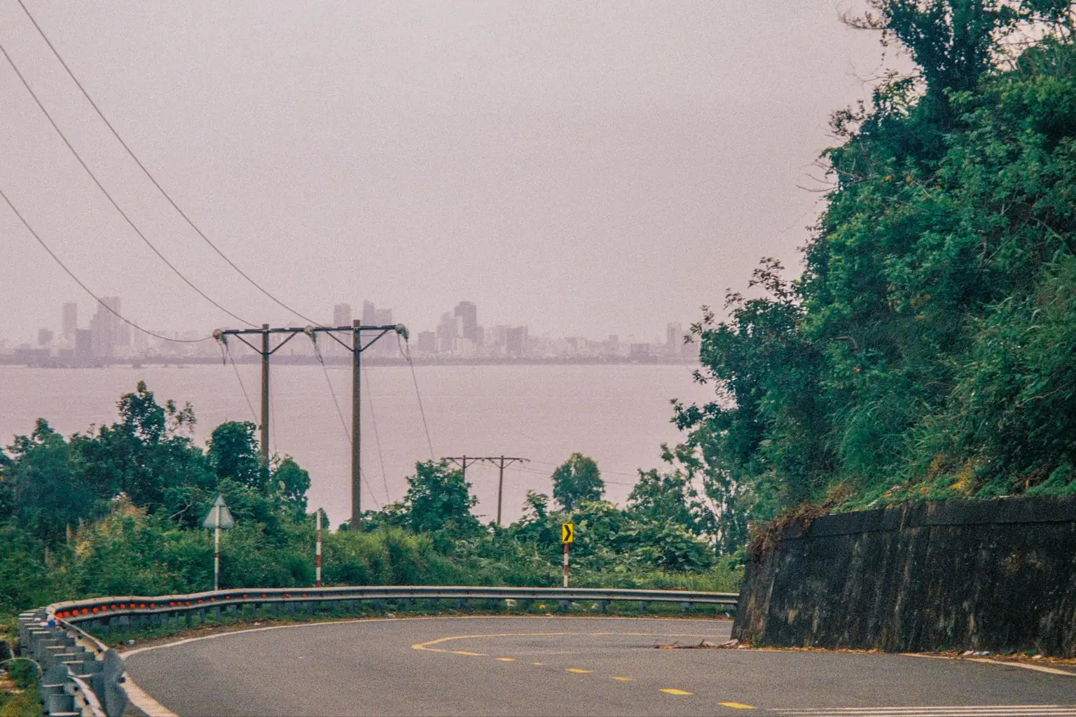 A curve of a mountain road looking out over a coastal city on an overcast day