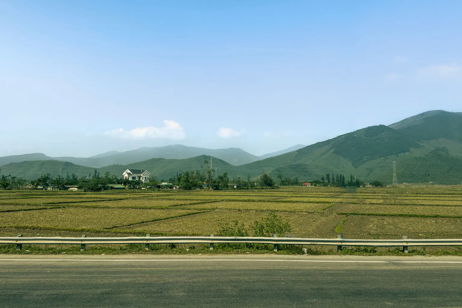 A road beside rice fields with green mountains in the distance under a clear sky